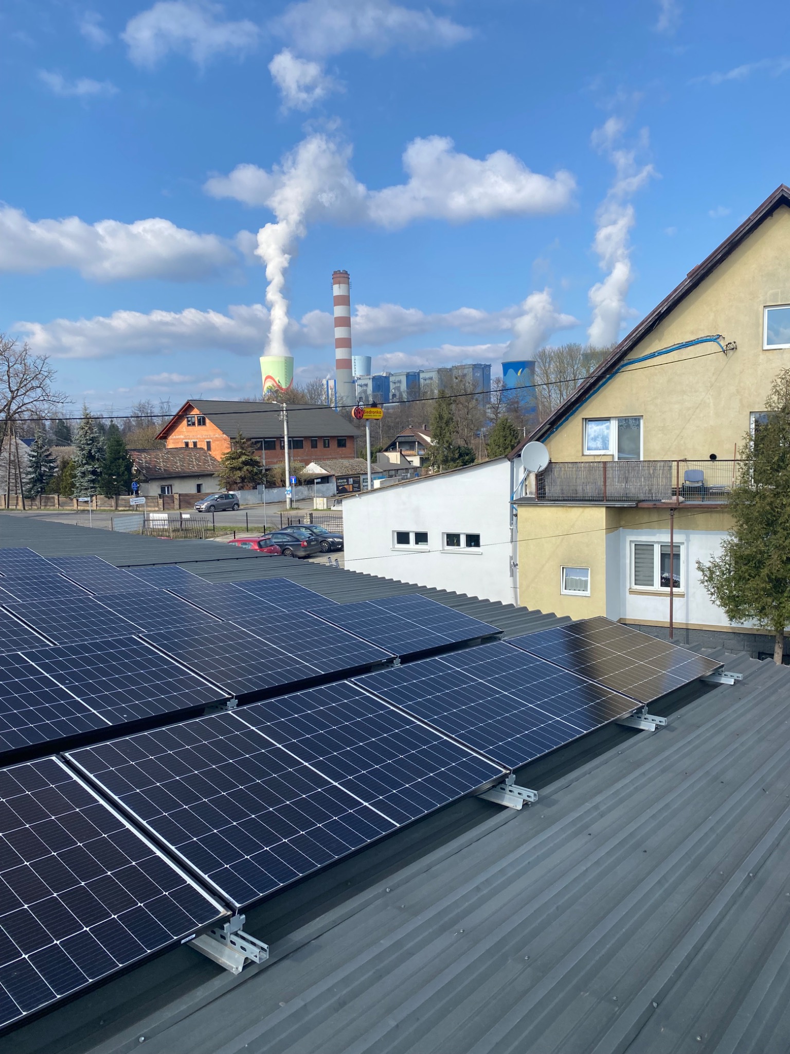 Panele fotowoltaiczne na dachu budynku mieszkalnego, w tle komin elektrowni i błękitne niebo z chmurami.