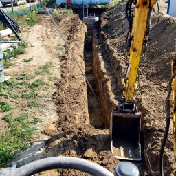 Michał Abel - Wąski wykop w ziemi, z widocznym betonowym kręgiem w głębi, wykonany koparką. Widok z kabiny operatora, w tle kontener biurowy i pojazdy.