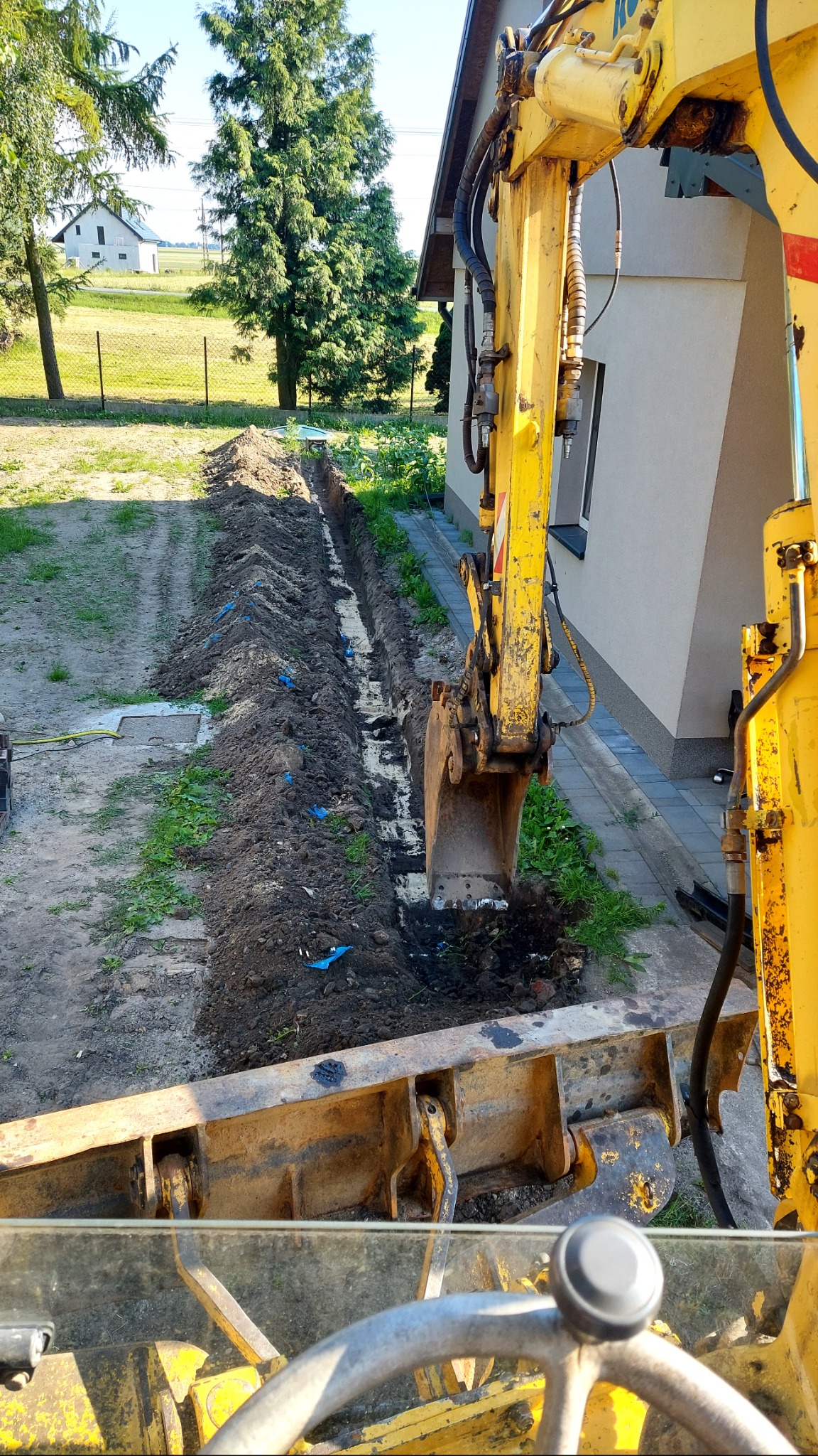 Żółty, używany sprzęt do wykopów podczas pracy nad wąskim wykopem w ziemi, prawdopodobnie pod instalacje, z widokiem na budynek i zieleń w tle.