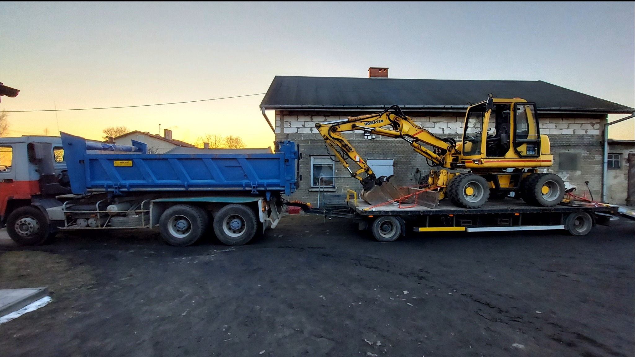 Ciężarówka z niebieską skrzynią ładunkową ciągnie lawetę z żółtą koparką Komatsu na tle ceglanego budynku i zachodzącego słońca.