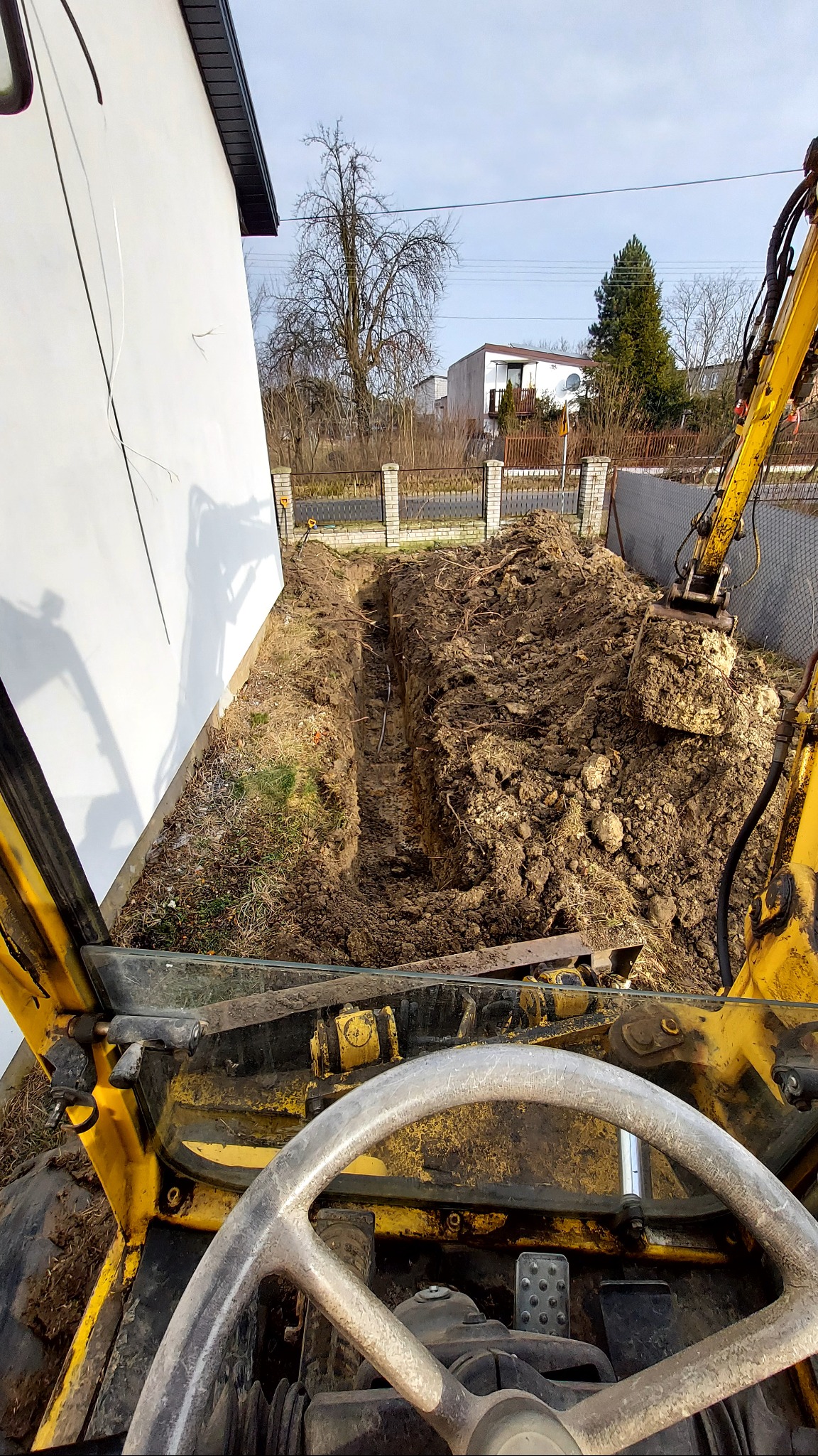 Wykop pod fundamenty domu jednorodzinnego, widok z kabiny koparki. W tle ogrodzenie i zabudowa. Ciemna ziemia, wykopana wąska bruzda wzdłuż ściany budynku.