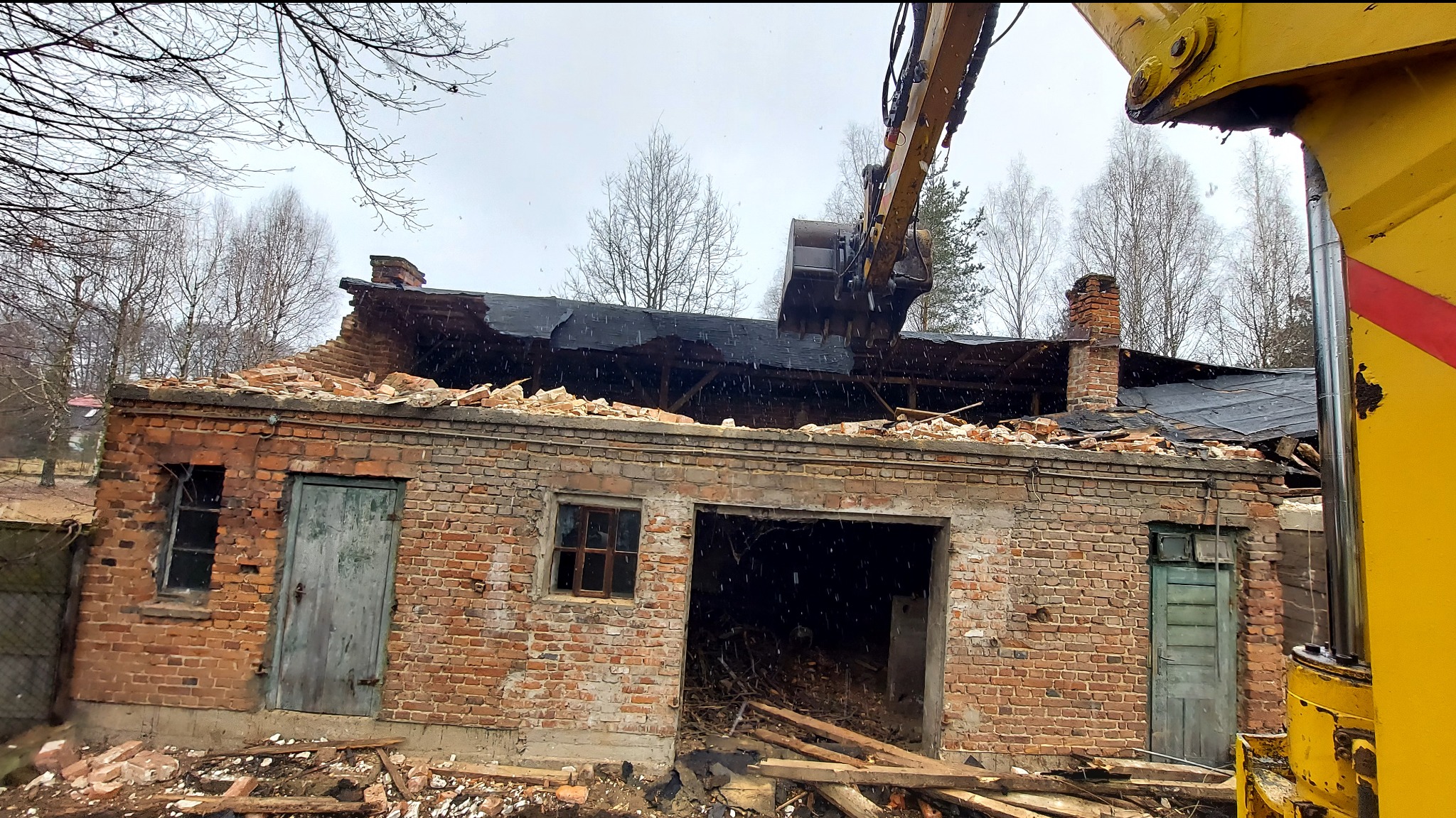 Ceglany budynek w trakcie rozbiórki przez koparkę z łyżką. Widoczne uszkodzenia dachu i ścian. Częściowo zburzony komin. Elementy żółtej maszyny na pierwszym planie.