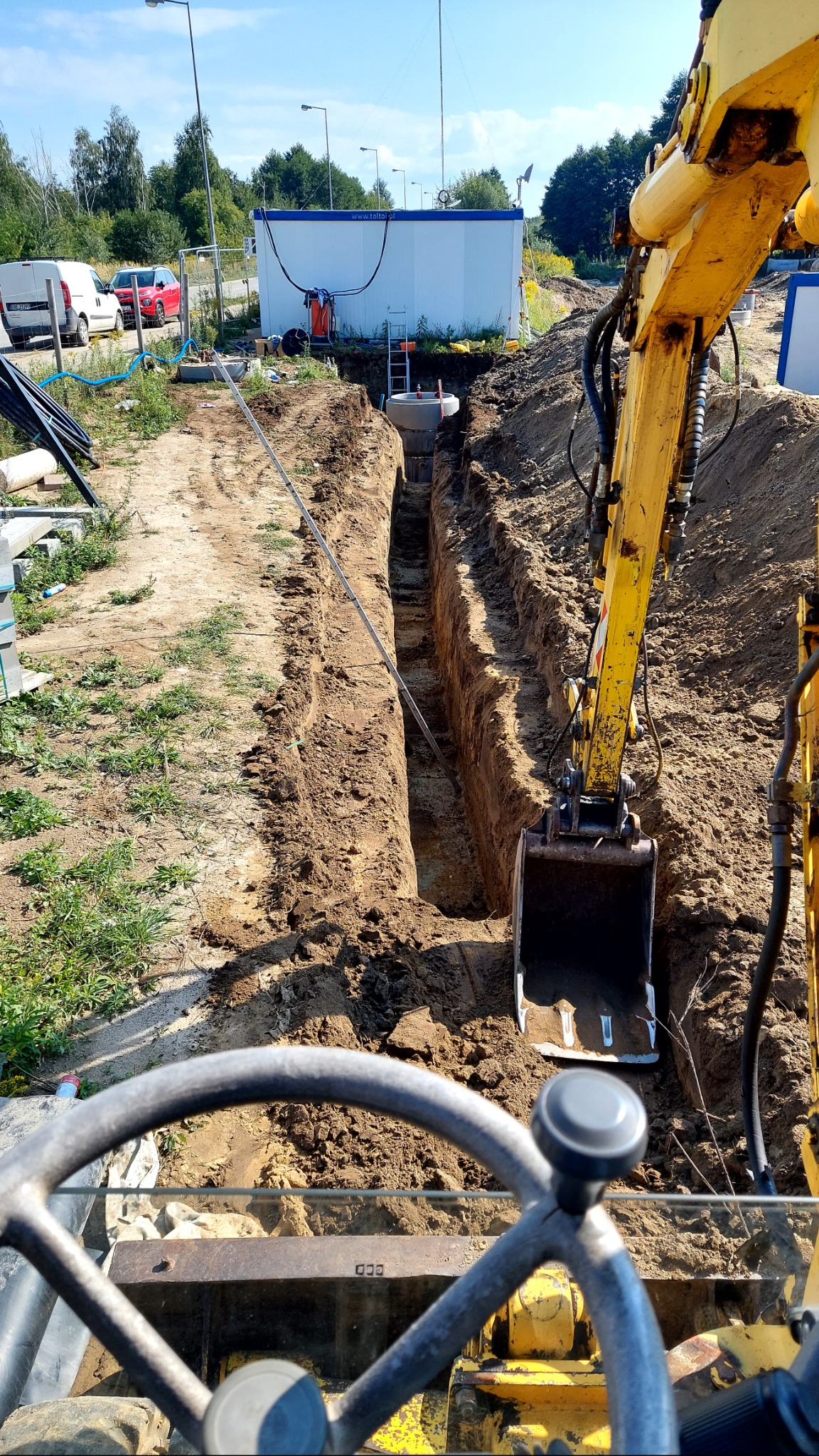 Wąski wykop w ziemi, z widocznym betonowym kręgiem w głębi, wykonany koparką. Widok z kabiny operatora, w tle kontener biurowy i pojazdy.