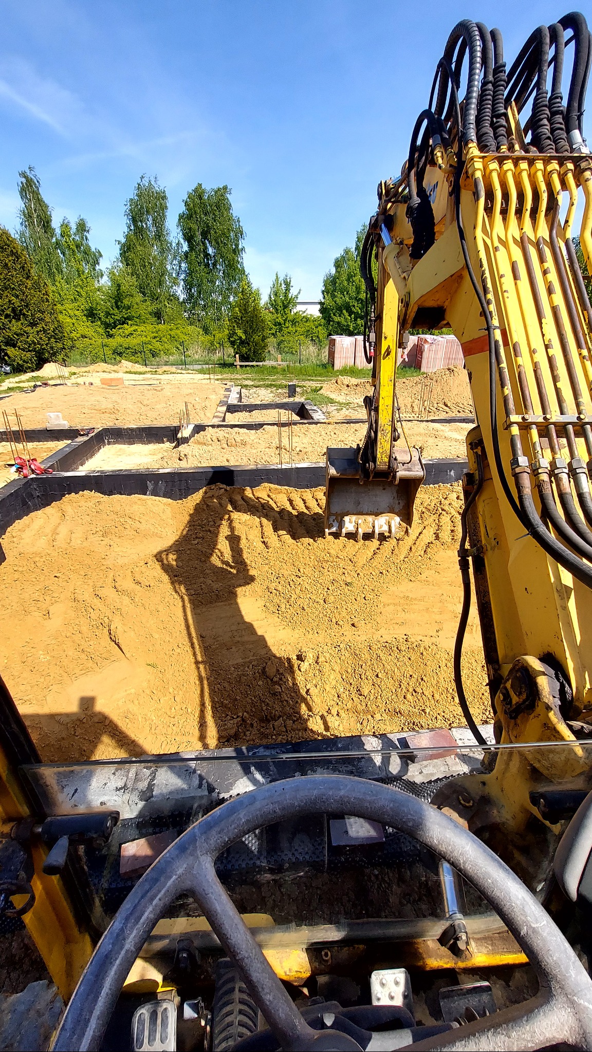Żółta koparka w trakcie wykopu pod fundamenty, widok z kabiny operatora, zarys fundamentów zbrojonych, błękitne niebo w tle.