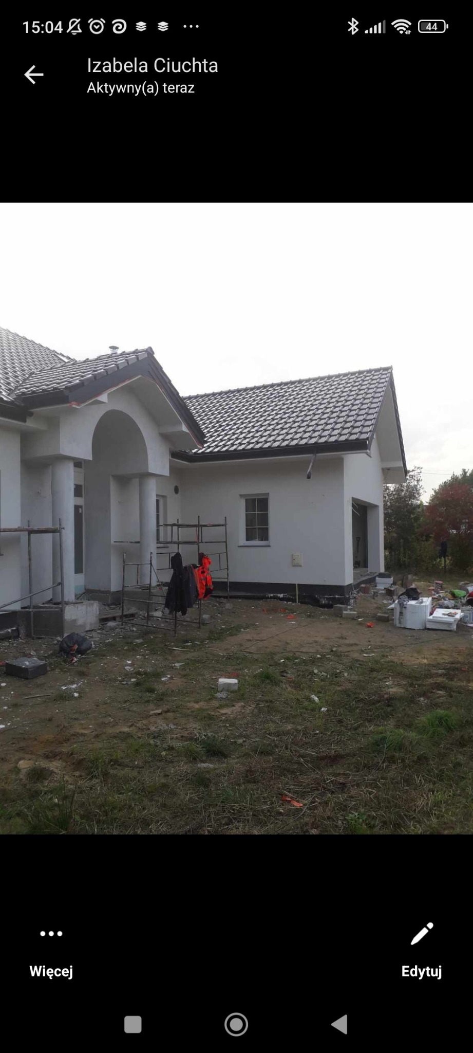 Niedokończony dom jednorodzinny z białymi ścianami i ciemną dachówką, widoczne rusztowania i materiały budowlane na trawniku, etap prac wykończeniowych.