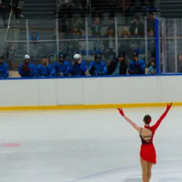 Kobieta w czerwonej sukience i rękawicach na lodowisku, z podniesionymi rękami, tyłem do widza, w tle siedząca publiczność i zawodnicy w niebieskich strojach.