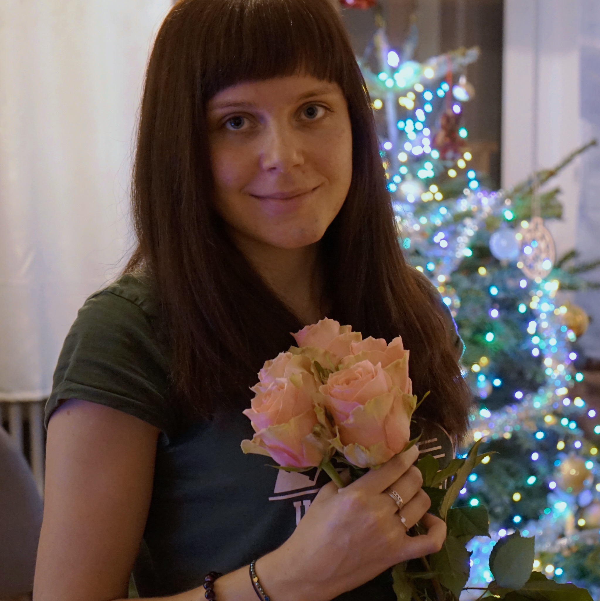 Portret kobiety z ciemnymi włosami i grzywką, trzymającej bukiet jasnoróżowych róż na tle rozświetlonej choinki.