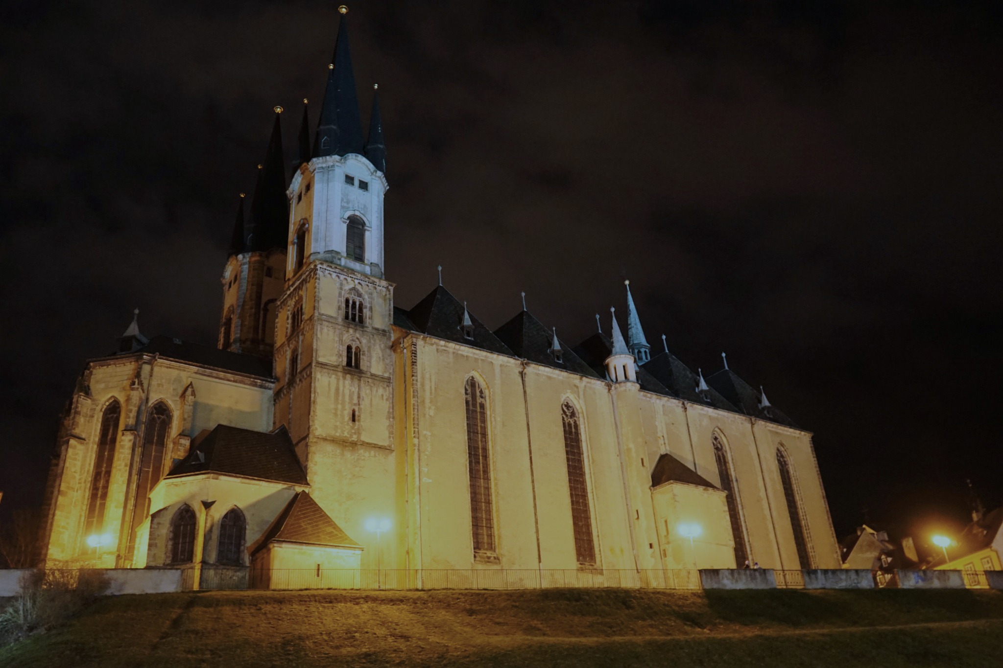 Oświetlony nocą gotycki kościół z wysokimi wieżami i iglicami, widoczny z dołu, na tle ciemnego nieba.