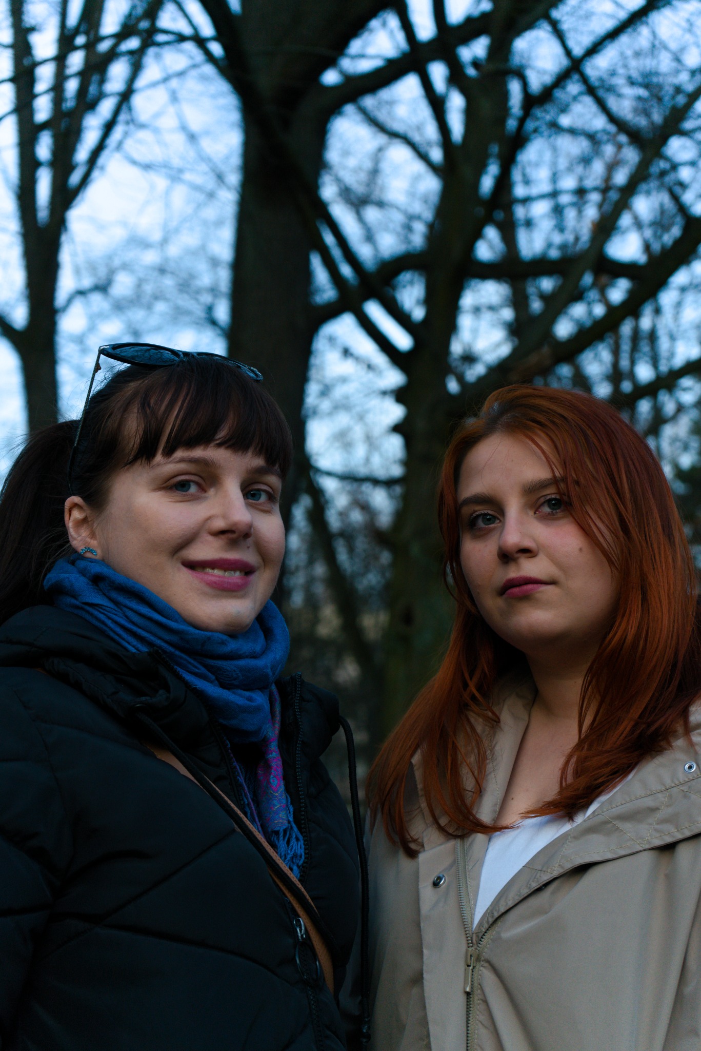 Dwie kobiety pozują na tle drzew, jedna uśmiechnięta w czarnej kurtce i niebieskim szalu, druga z rudymi włosami w beżowej kurtce.