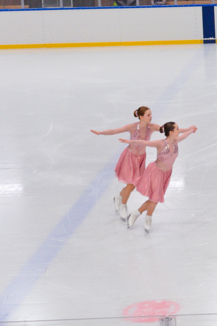 Dwie młode kobiety w różowych sukienkach, wykonujące synchroniczne figury na lodowisku, z rozpostartymi ramionami i uniesionymi głowami.