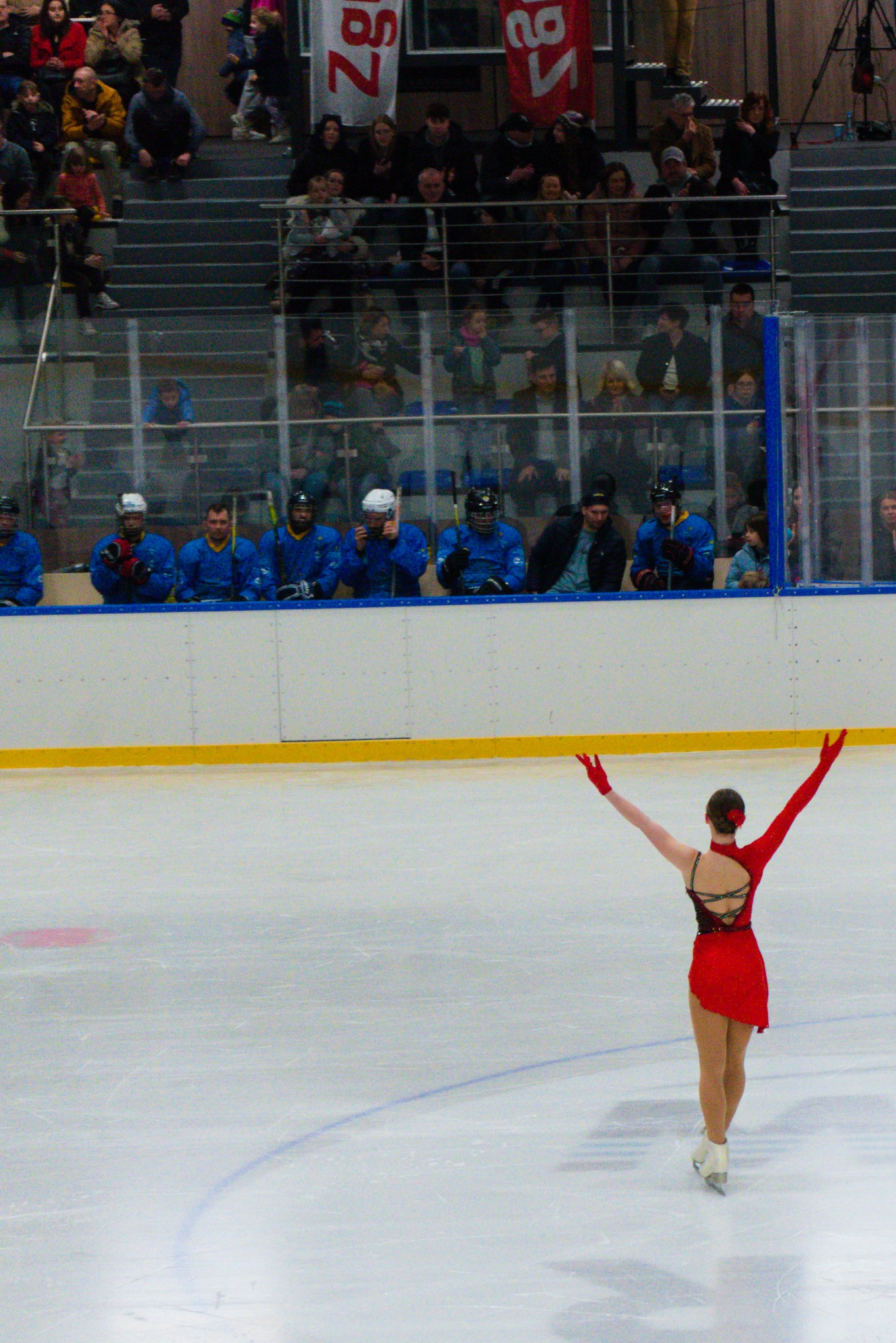 Kobieta w czerwonej sukience i rękawicach na lodowisku, z podniesionymi rękami, tyłem do widza, w tle siedząca publiczność i zawodnicy w niebieskich strojach.