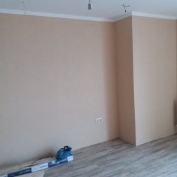 VELVET MIX DECORBUD Yurii Raba - Pomalowane na beżowo ściany w pustym pokoju z drewnopodobną podłogą, białym grzejnikiem i listwami przypodłogowymi. Na podłodze leżą panele i niebieska torba narzędziowa.