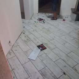 VELVET MIX DECORBUD Yurii Raba - Układanie podłogi z płytek imitujących drewno w Warszawie, widoczne krzyżyki dystansowe i narzędzia glazurnicze, osoba przechodząca w tle.