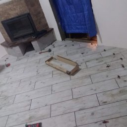 VELVET MIX DECORBUD Yurii Raba - Układanie podłogi z płytek imitujących drewno w pomieszczeniu z kominkiem, widoczne krzyżyki dystansowe i karton po płytkach.