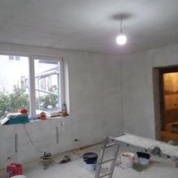 VELVET MIX DECORBUD Yurii Raba - Pokój w trakcie przygotowania do malowania, ściany pokryte gładzią, widoczne narzędzia, drabina, okno z widokiem na zieleń na zewnątrz.