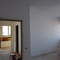 VELVET MIX DECORBUD Yurii Raba - Wnętrze w trakcie remontu: świeżo pomalowane białe ściany, sufit z listwą dekoracyjną, widok na sąsiednie pomieszczenie z drewnianą boazerią, worki z gładzią na podłodze.