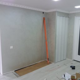 VELVET MIX DECORBUD Yurii Raba - Świeżo pomalowane ściany w odcieniach szarości z dekoracyjną sztukaterią pod sufitem, poziomica oparta o ścianę, przygotowane podłoże pod nową podłogę, widoczne narzędzia i akcesoria malarskie.