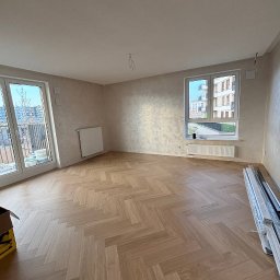 VELVET MIX DECORBUD Yurii Raba - Pusty pokój z parkietem ułożonym w jodełkę, ściany wykończone dekoracyjnym tynkiem, widoczne okno z widokiem na budynek mieszkalny i częściowo widoczne elementy grzejników.