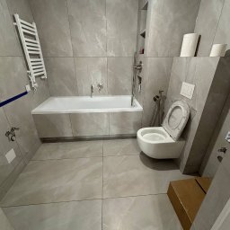 VELVET MIX DECORBUD Yurii Raba - Nowoczesna łazienka z szarymi płytkami imitującymi marmur, wiszącą toaletą z deską w folii ochronnej, zabudowaną wanną i grzejnikiem drabinkowym. Na podłodze duże płytki w odcieniach szarości.