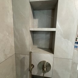 VELVET MIX DECORBUD Yurii Raba - Wykończona wnęka prysznicowa z półkami, wyłożona płytkami imitującymi marmur, z zamontowaną baterią prysznicową w kolorze szczotkowanego niklu.