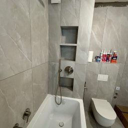 VELVET MIX DECORBUD Yurii Raba - Nowoczesna łazienka w trakcie wykańczania: szare marmurowe płytki, biała wanna i toaleta, półki w zabudowie, widoczne materiały wykończeniowe i niezakończone prace przy suficie.