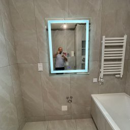 VELVET MIX DECORBUD Yurii Raba - Jasnoszara łazienka z dużymi płytkami, podświetlanym lustrem, grzejnikiem drabinkowym i wanną, widoczne ślady po fugowaniu pod sufitem, odbicie osoby robiącej zdjęcie w lustrze.