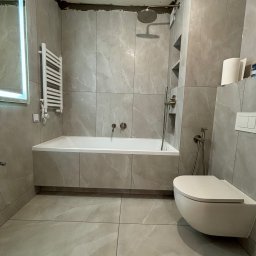 VELVET MIX DECORBUD Yurii Raba - Wykończona łazienka w szarych płytkach imitujących marmur, z podwieszaną toaletą, zabudowaną wanną, grzejnikiem drabinkowym i oświetlonym lustrem, widoczne niedokończone prace wykończeniowe...