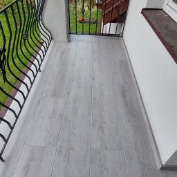 VELVET MIX DECORBUD Yurii Raba - Balkon z szarą podłogą imitującą drewno, czarna, metalowa balustrada z ozdobnymi elementami, widok na zieleń w tle.