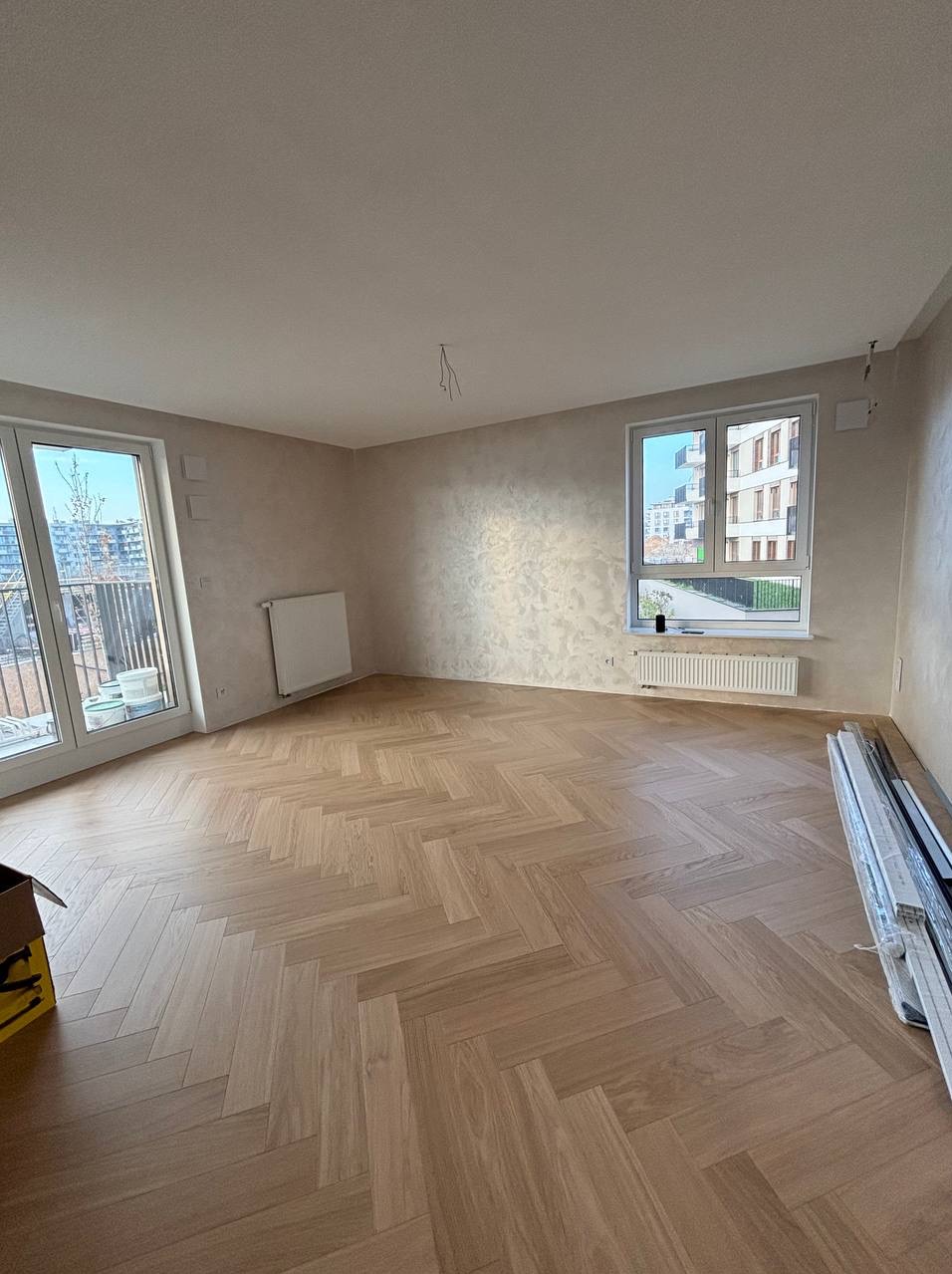 Pusty pokój z parkietem ułożonym w jodełkę, ściany wykończone dekoracyjnym tynkiem, widoczne okno z widokiem na budynek mieszkalny i częściowo widoczne elementy grzejników.