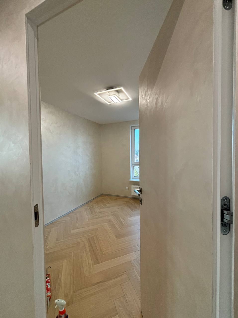 Wykończone wnętrze pokoju z parkietem w jodełkę, ściany pokryte dekoracyjną szpachlą w odcieniach beżu, widok przez otwarte drzwi, w tle okno z widokiem na zewnątrz.