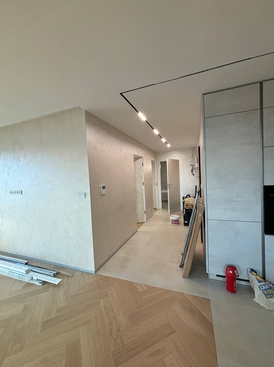 Korytarz w trakcie prac wykończeniowych: położona podłoga w jodełkę łączy się z płytkami, ściany z efektem dekoracyjnym, widoczne listwy przypodłogowe, puszka z farbą i narzędzia.