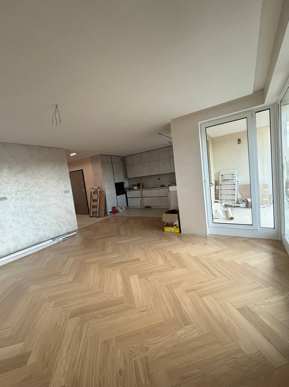 Jasny parkiet ułożony we wzór jodełki w przestronnym, częściowo wykończonym wnętrzu z widoczną kuchnią i świeżo pomalowaną ścianą.