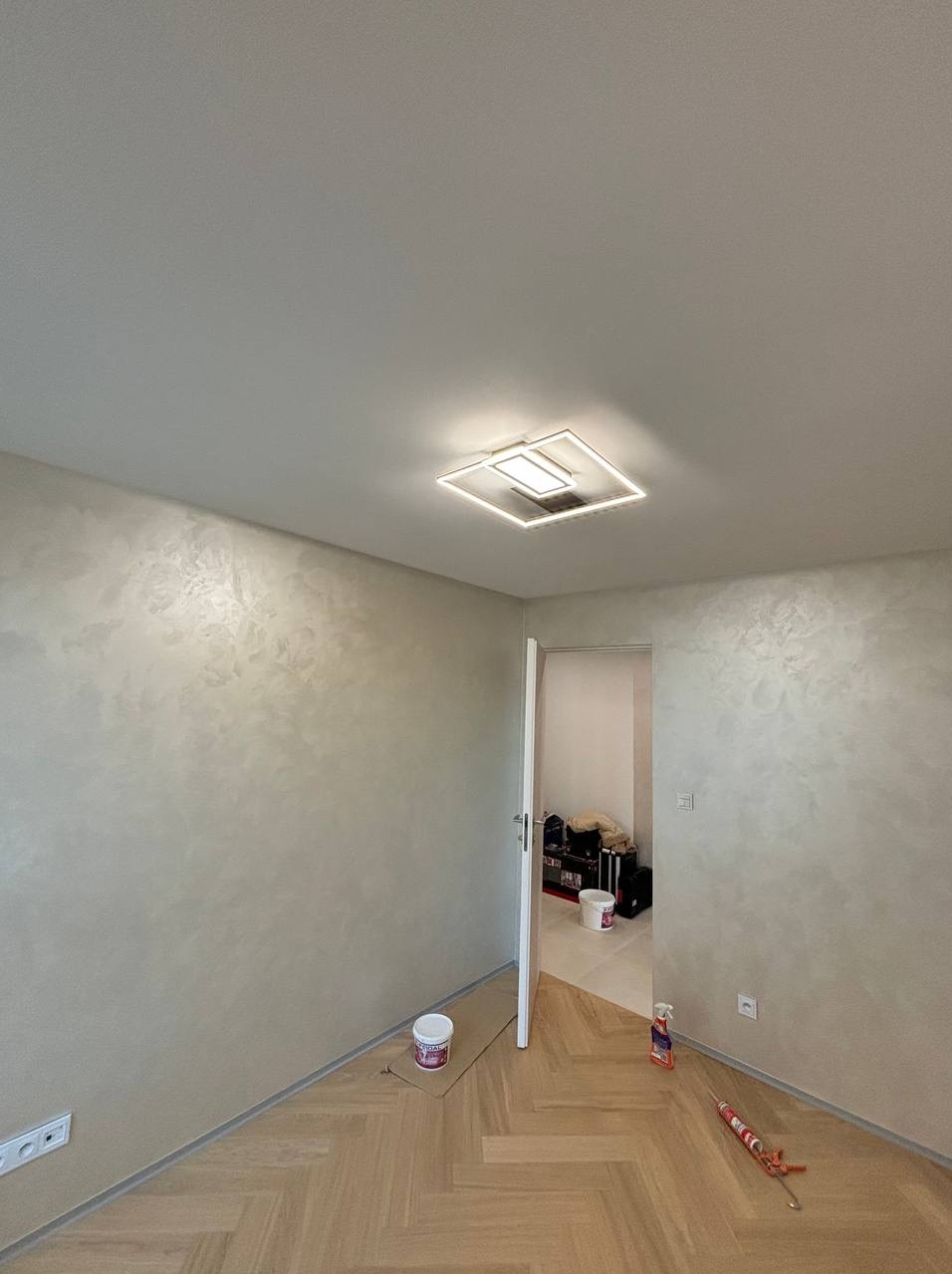 Pokój po szpachlowaniu i malowaniu dekoracyjnym, widoczny parkiet w jodełkę, nowoczesna lampa sufitowa i otwarte drzwi do kolejnego pomieszczenia, gdzie widać narzędzia i wiadro.