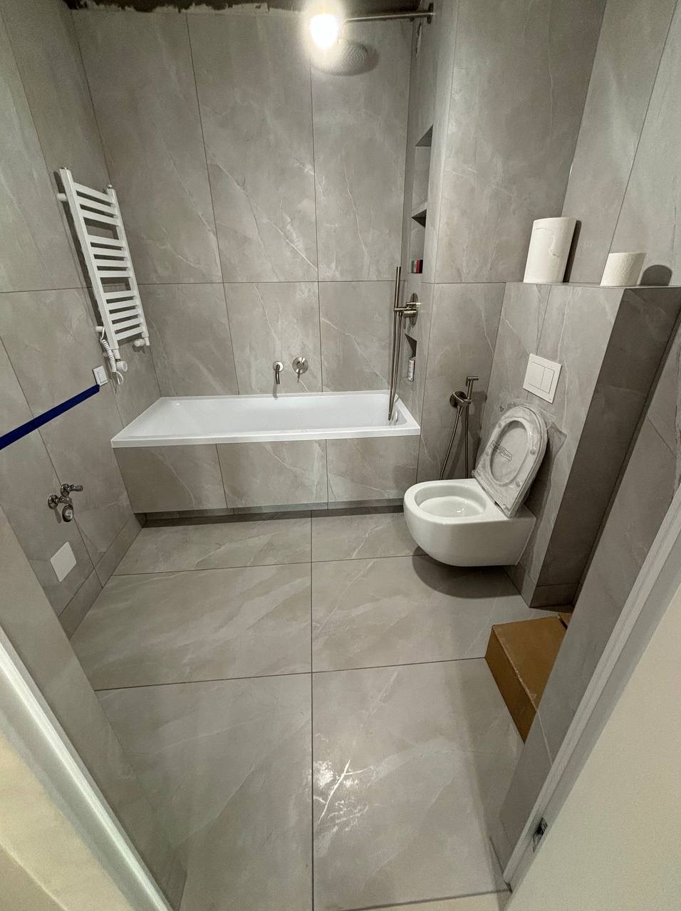 Nowoczesna łazienka wykończona płytkami imitującymi marmur, z wiszącą toaletą, wanną i grzejnikiem drabinkowym, oświetlona górnym światłem.