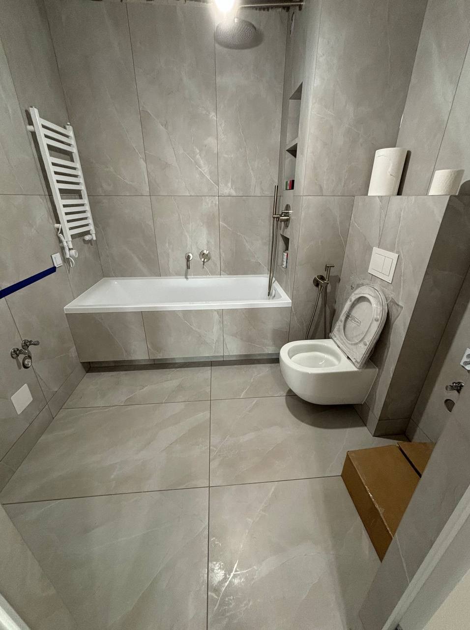 Nowoczesna łazienka z szarymi płytkami imitującymi marmur, wiszącą toaletą z deską w folii ochronnej, zabudowaną wanną i grzejnikiem drabinkowym. Na podłodze duże płytki w odcieniach szarości.