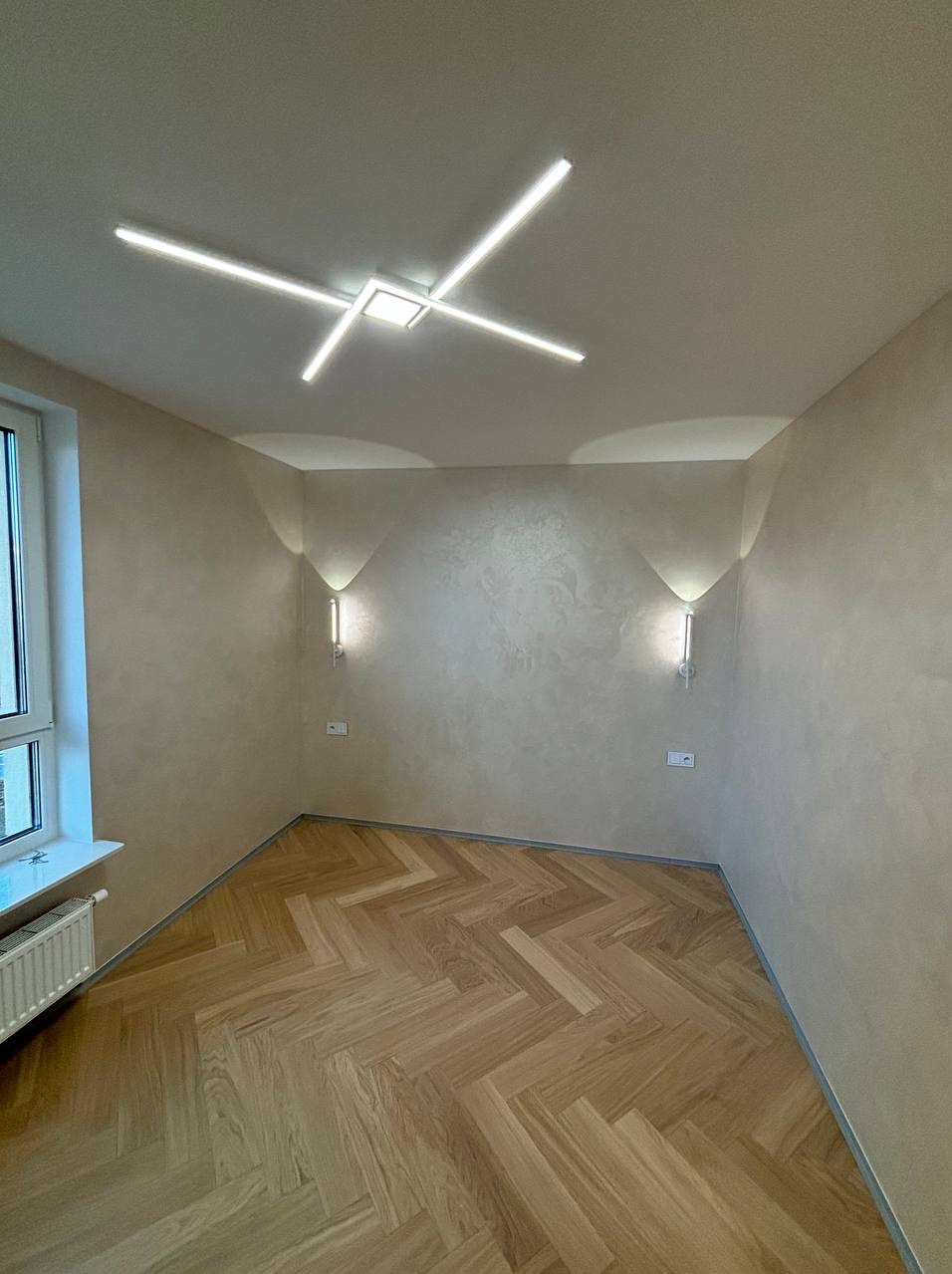 Pokój z parkietem ułożonym w jodełkę, ścianami wykończonymi dekoracyjnym tynkiem, nowoczesnym oświetleniem sufitowym w kształcie krzyża i minimalistycznymi kinkietami ściennymi.