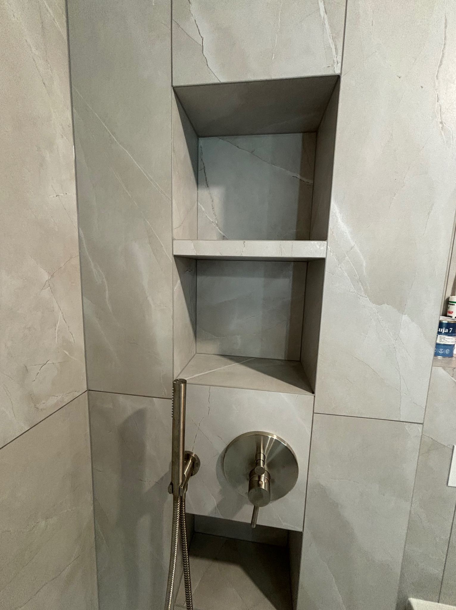 Wykończona wnęka prysznicowa z półkami, wyłożona płytkami imitującymi marmur, z zamontowaną baterią prysznicową w kolorze szczotkowanego niklu.