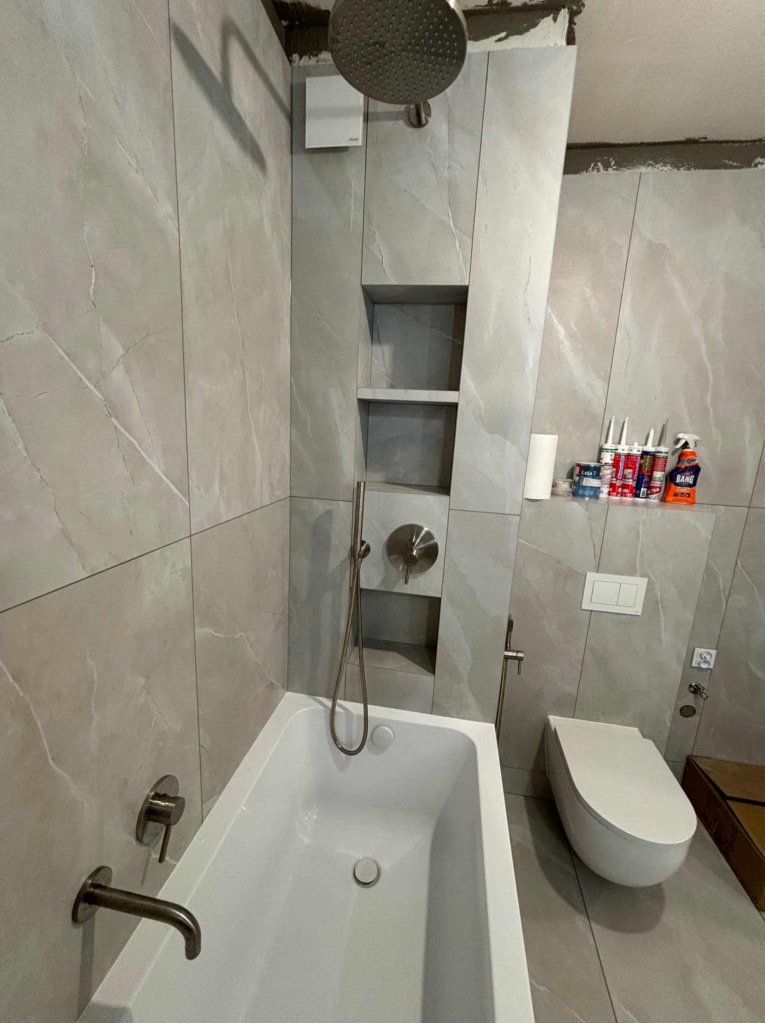 Nowoczesna łazienka w trakcie wykańczania: szare marmurowe płytki, biała wanna i toaleta, półki w zabudowie, widoczne materiały wykończeniowe i niezakończone prace przy suficie.