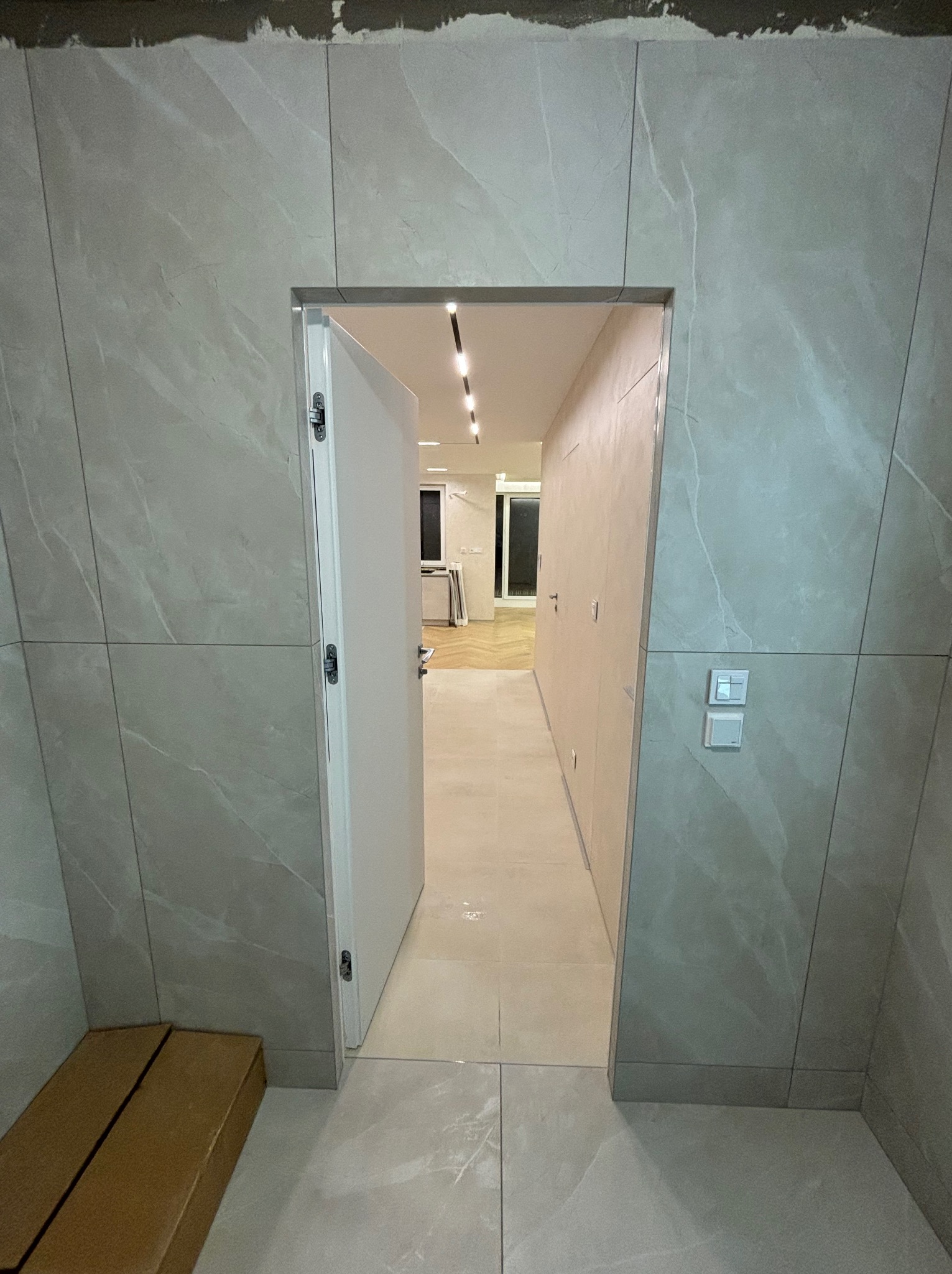 Wykończone wnętrze z otwartymi, białymi drzwiami, widoczne szare, wielkoformatowe płytki imitujące marmur na ścianach i podłodze, oraz fragment kartonów na podłodze.