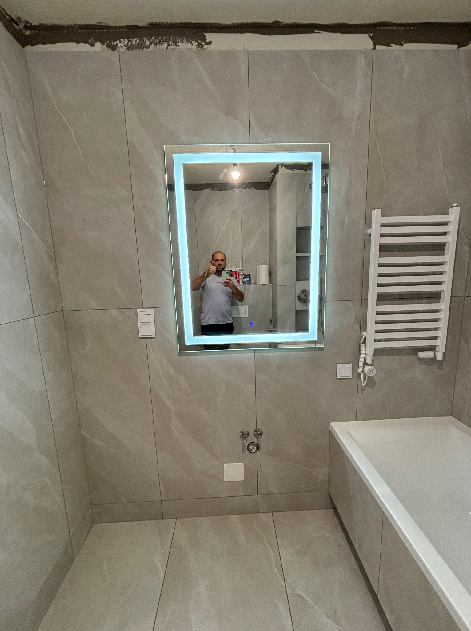 Jasnoszara łazienka z dużymi płytkami, podświetlanym lustrem, grzejnikiem drabinkowym i wanną, widoczne ślady po fugowaniu pod sufitem, odbicie osoby robiącej zdjęcie w lustrze.