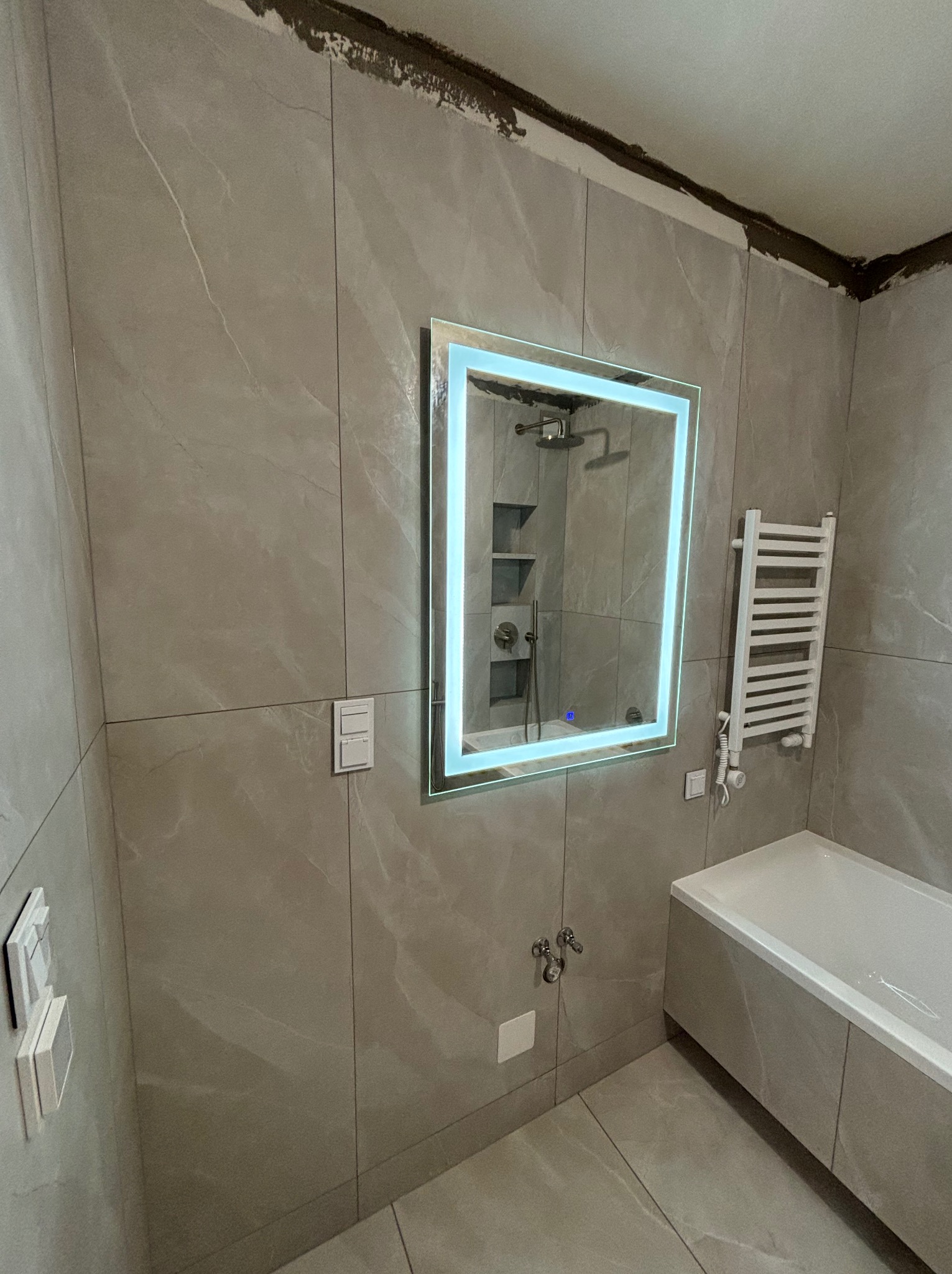 Wykończona łazienka z płytkami imitującymi marmur, podświetlane lustro z widocznym prysznicem w odbiciu, grzejnik drabinkowy i biała wanna. Na górze ściany widoczne ślady po kleju lub zaprawie.