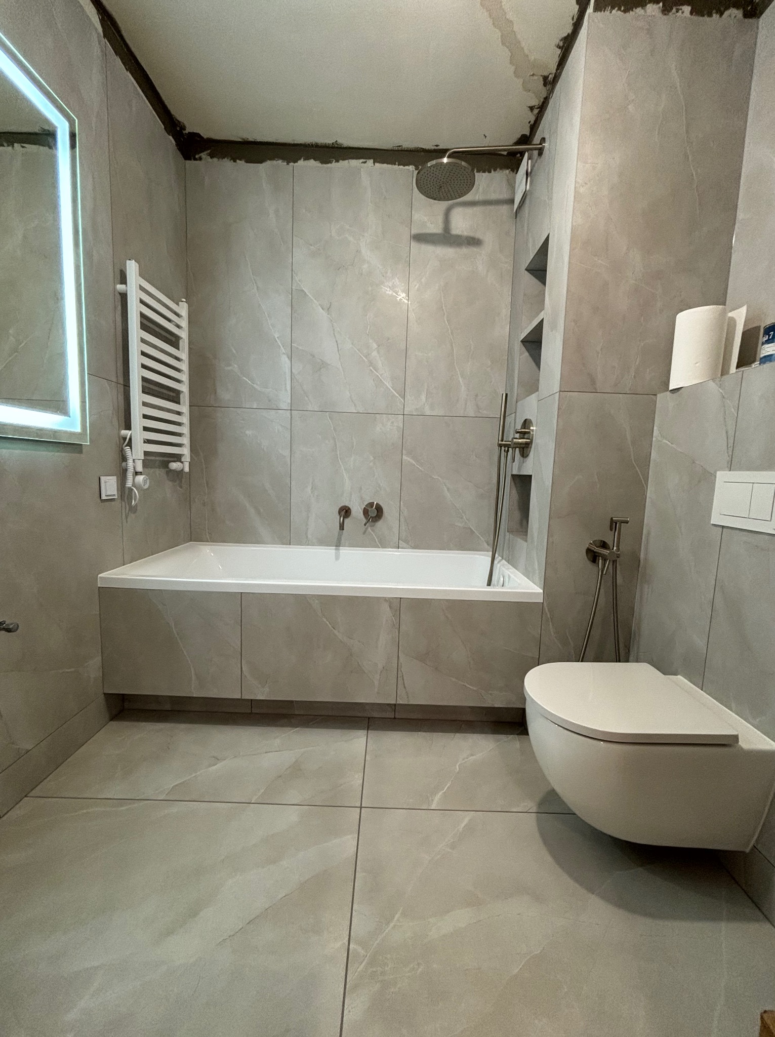 Wykończona łazienka w szarych płytkach imitujących marmur, z podwieszaną toaletą, zabudowaną wanną, grzejnikiem drabinkowym i oświetlonym lustrem, widoczne niedokończone prace wykończeniowe...