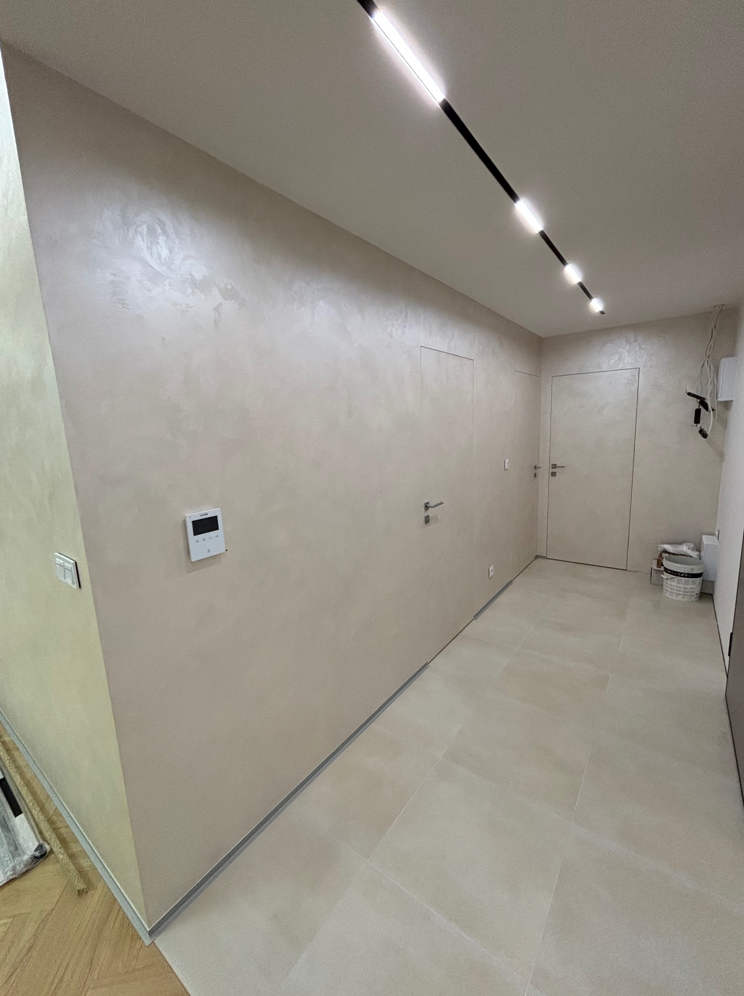 Korytarz z beżowymi płytkami podłogowymi, ścianami pokrytymi dekoracyjnym tynkiem w odcieniach beżu z subtelnym połyskiem, wbudowanymi drzwiami bez listew oraz nowoczesnym oświetleniem liniowym...