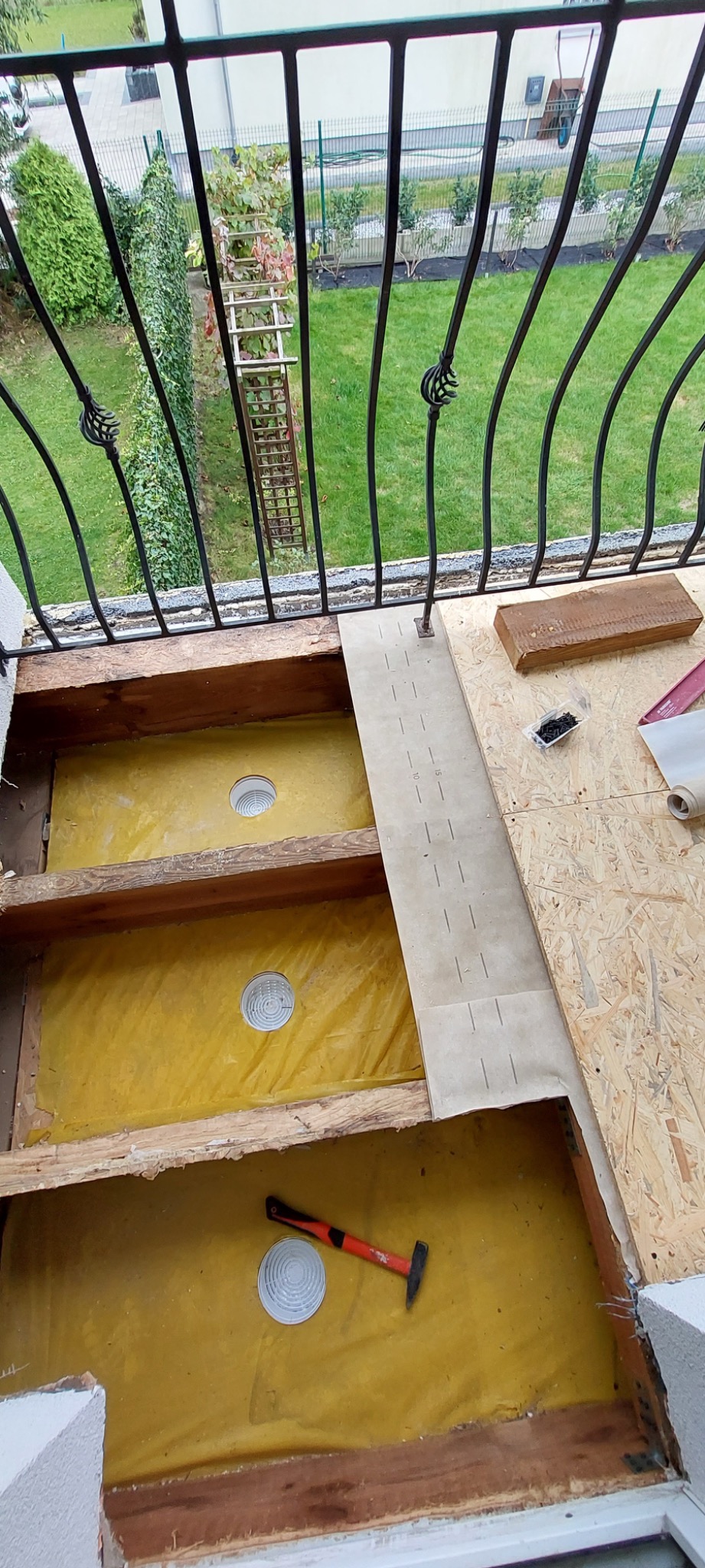 Balkon w trakcie izolacji podłogi, widoczne drewniane legary, żółta folia izolacyjna, otwory wentylacyjne, płyta OSB, listwa pomiarowa i młotek z czerwonym trzonkiem.