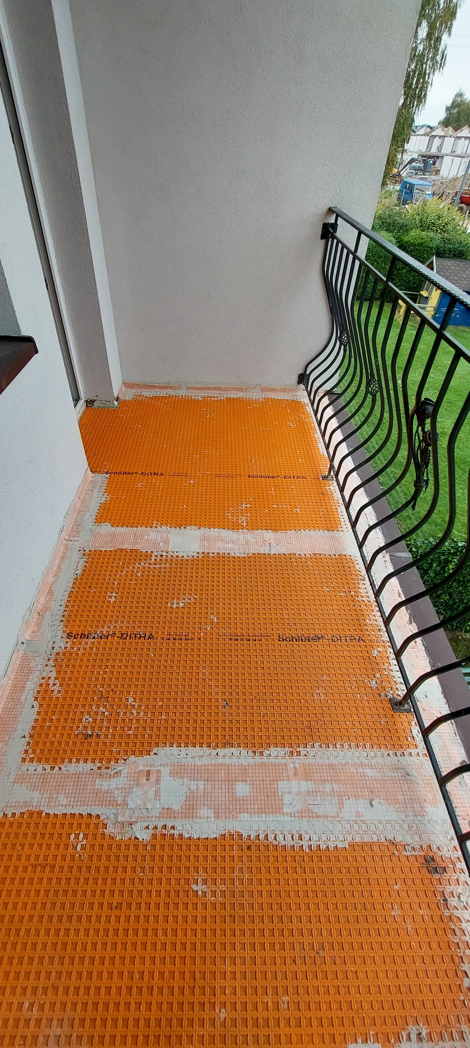Pomarańczowa mata drenażowa Schluter-DITRA ułożona na balkonie, widoczne ślady zaprawy klejowej, czarna metalowa balustrada z ozdobnymi zakończeniami, widok na zieleń w tle.