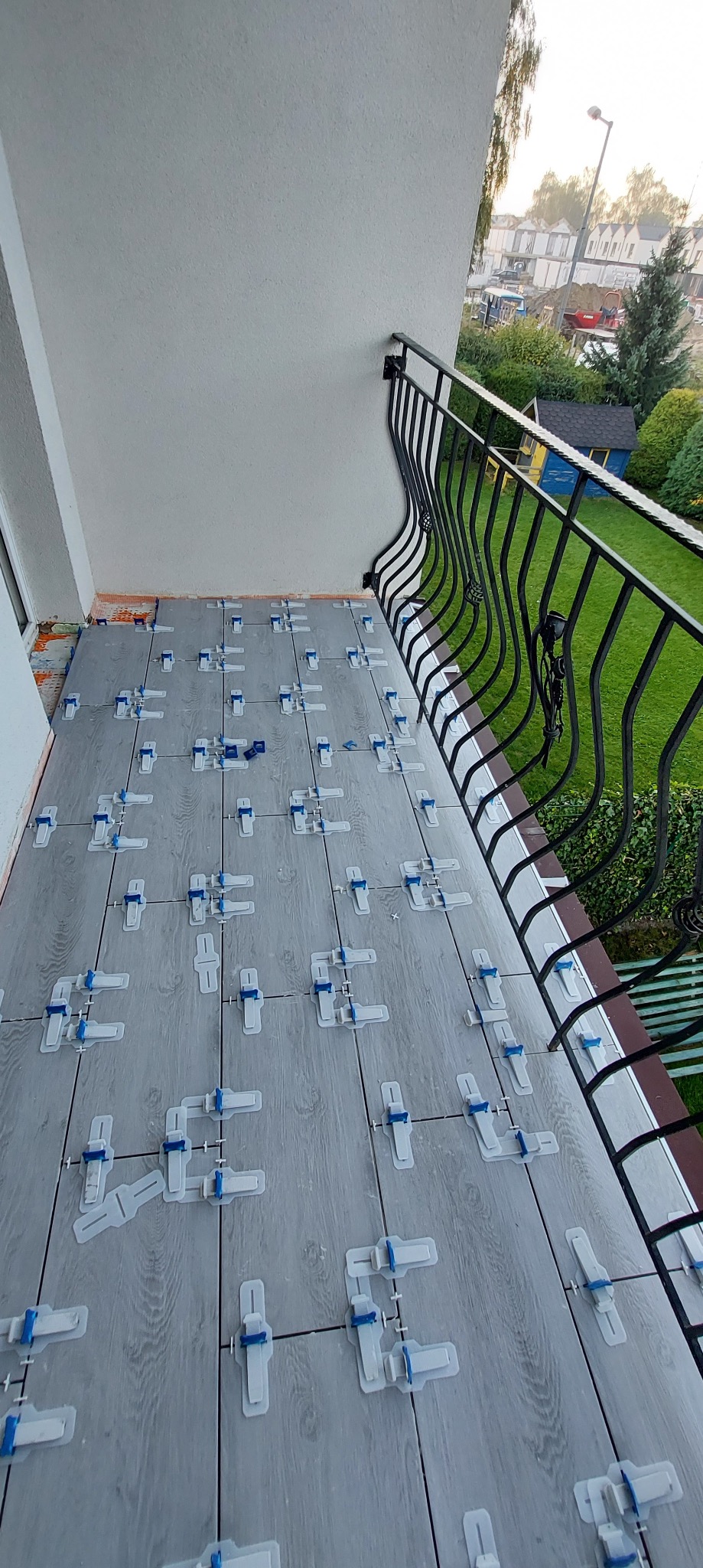 Szare płytki imitujące drewno na balkonie, z systemem poziomowania płytek (klipsy i kliny), widok na zewnątrz z ozdobną, czarną balustradą.