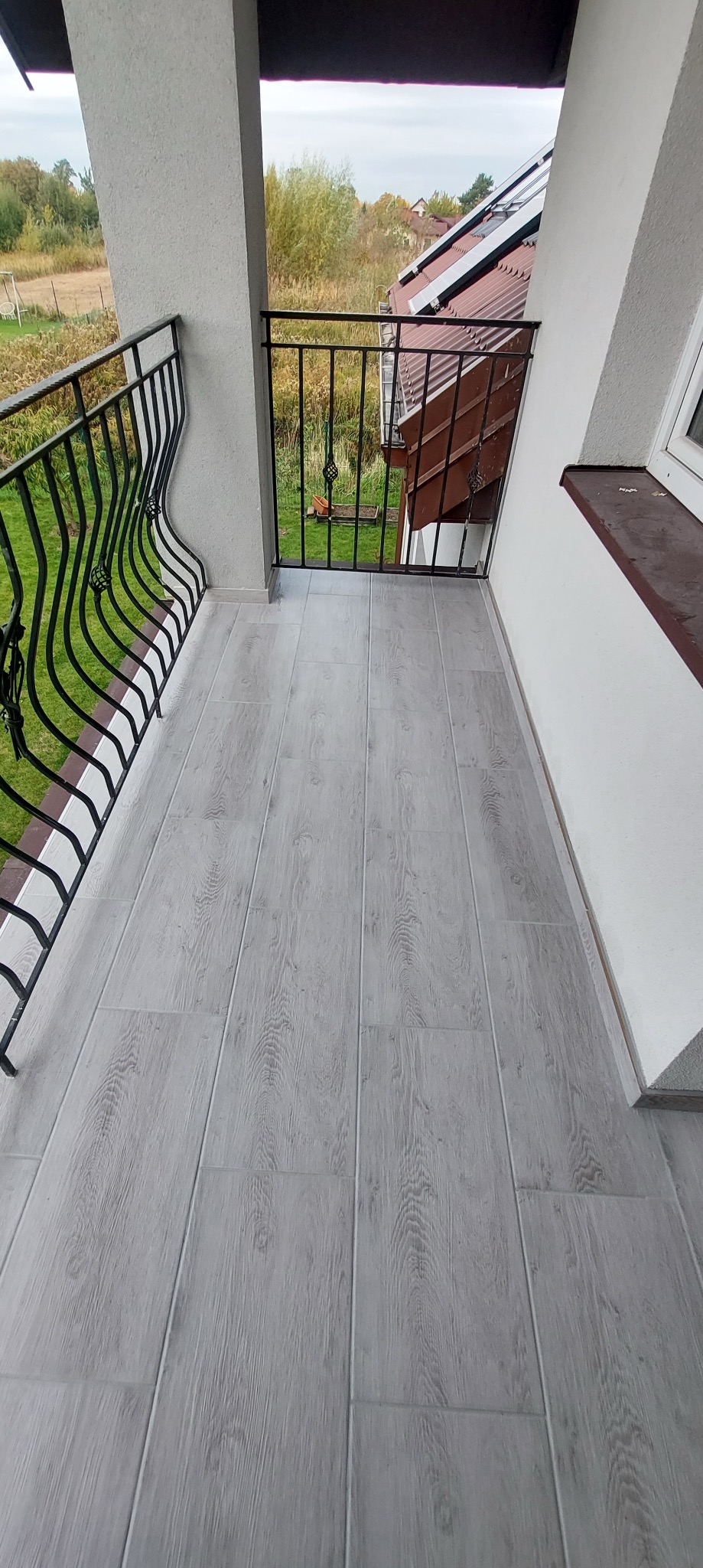 Balkon z szarą podłogą imitującą drewno, czarna, metalowa balustrada z ozdobnymi elementami, widok na zieleń w tle.