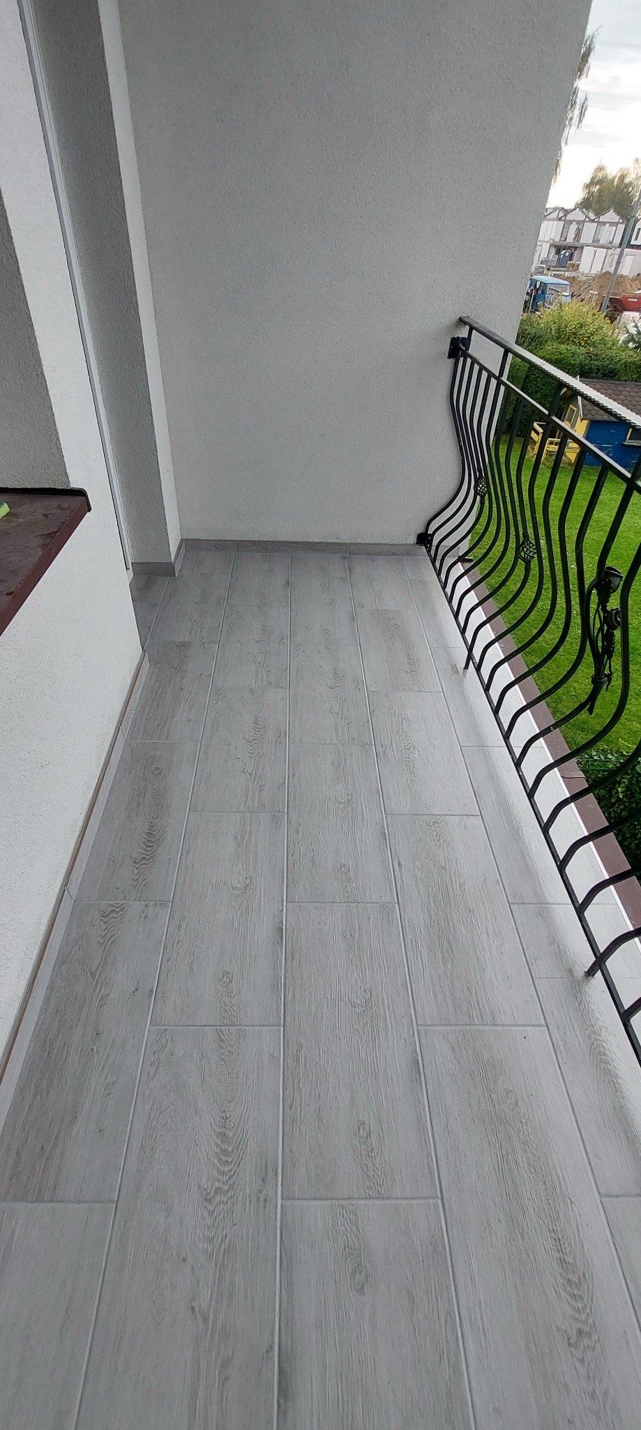 Wyłożony płytkami balkon z ozdobną, metalową balustradą i widokiem na zieleń.