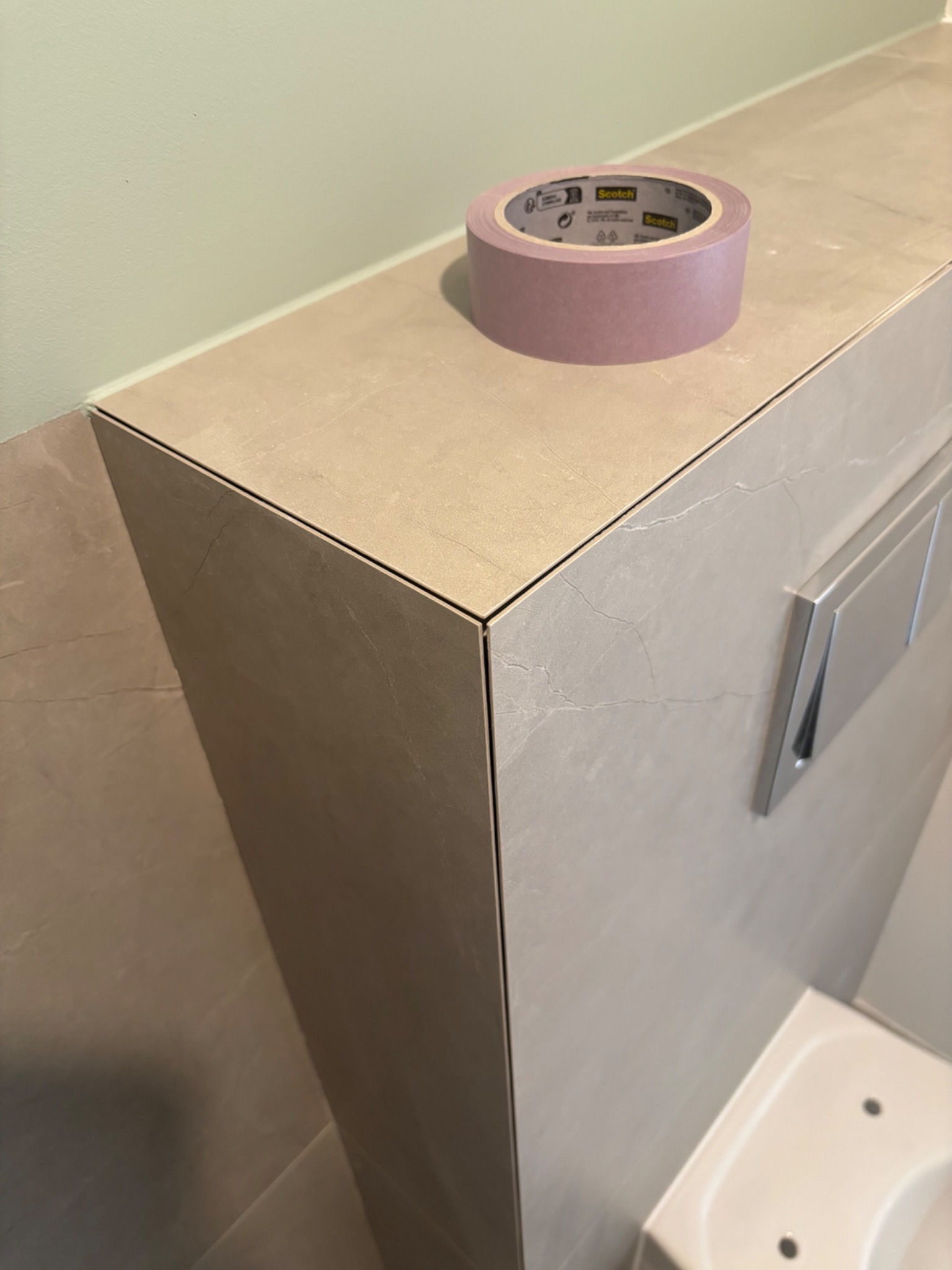 Fragment łazienki z obudową stelaża WC wykończoną płytkami imitującymi marmur, na górze leży rolka taśmy malarskiej Scotch, widoczny fragment toalety