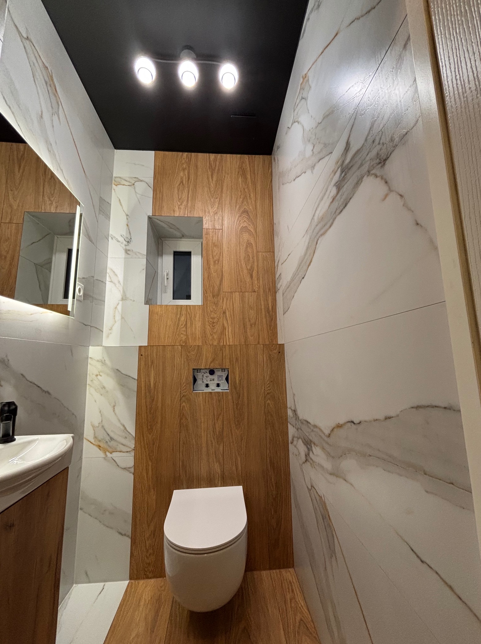 Nowoczesna łazienka z białymi marmurkowymi płytkami i drewnianymi panelami, wisząca toaleta, czarny sufit z trzema punktowymi lampami, podświetlane lustro i wnęka z oknem.